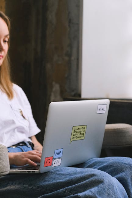 A close-up view of a laptop resting on a person's lap, situated outdoors in a garden or backyard. The laptop's cover displays several colorful stickers, including one with binary code, a PHP logo, and